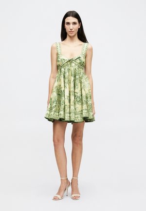 Γυναίκα με μακριά σκούρα μαλλιά που φορά ένα αμάνικο, κοντό φόρεμα με πράσινο μοτίβο και μπεζ ψηλοτάκουνα σανδάλια, στέκεται μπροστά σε σκηνοθετημένο φόντο.