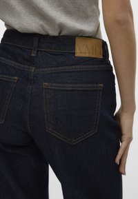 Jeans de mezclilla oscura con corte recto, dos bolsillos traseros y una etiqueta de cuero marrón con texto en relieve en la cintura.