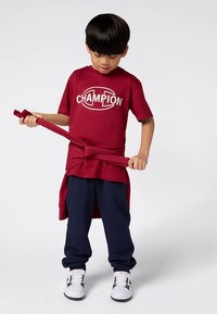 Punane puuvillane T-särk "CHAMPION" logoga, tumesinised dressipüksid ja valged tossud. Särk on vöökohalt sõlmega seotud.