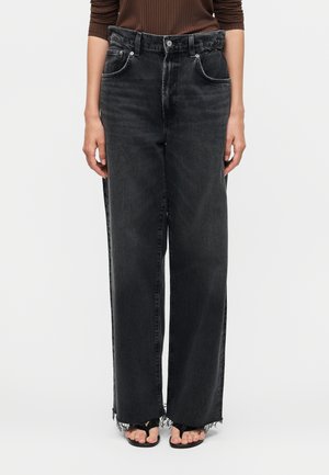 Person iført sort højtaljede vidde jeans med frynset kant og sorte åbne sandaler, kombineret med en brun ribbet top.