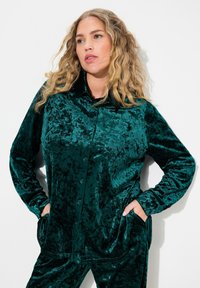 Camisa verde terciopelo de botones con mangas largas, dos bolsillos en el pecho y un cuello tipo camisa, que presenta una textura suave y brillante.
