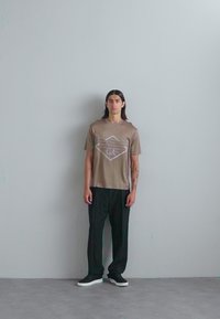 Brun T-skjorte med kort erm og en geometrisk grafikkdesign på brystet, kombinert med svarte bukser og sko. Subtil tekstur i stoffet.