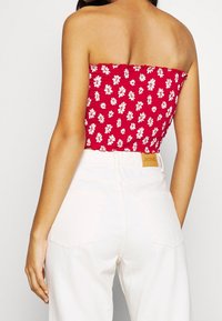 Žena v červeném květovaném topu bez ramínek a bílých kalhotách s vysokým pasem, zobrazená zezadu na jednobarevném pozadí.