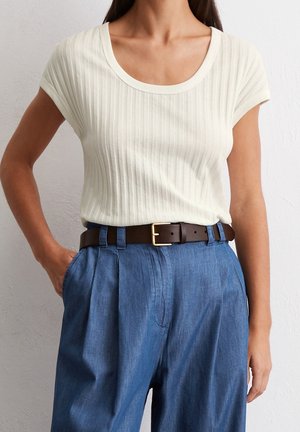 Frau trägt ein cremefarbenes, kurzärmeliges, geripptes Oberteil, das in hoch taillierte blaue, plissierte Hosen mit einem dunkelbraunen Gürtel gesteckt ist.