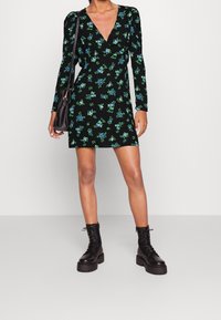 Robe noire à manches longues avec un profond décolleté en V, ornée de motifs floraux bleus et verts. Associée à des bottes noires à lacets épaisses.