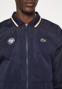 Man som bär en marinblå zip-up jacka med randig krage, Roland Garros Paris-logotyp på vänster bröst och grön Lacoste krokodil-logotyp på höger bröst.