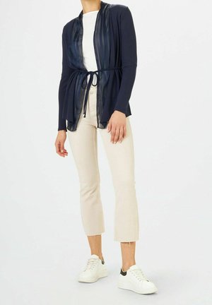 Frau trägt marineblauen Cardigan mit Gürtel über weißem Hemd, beige verkürzte Hose und weiße Turnschuhe vor schlichtem Hintergrund.