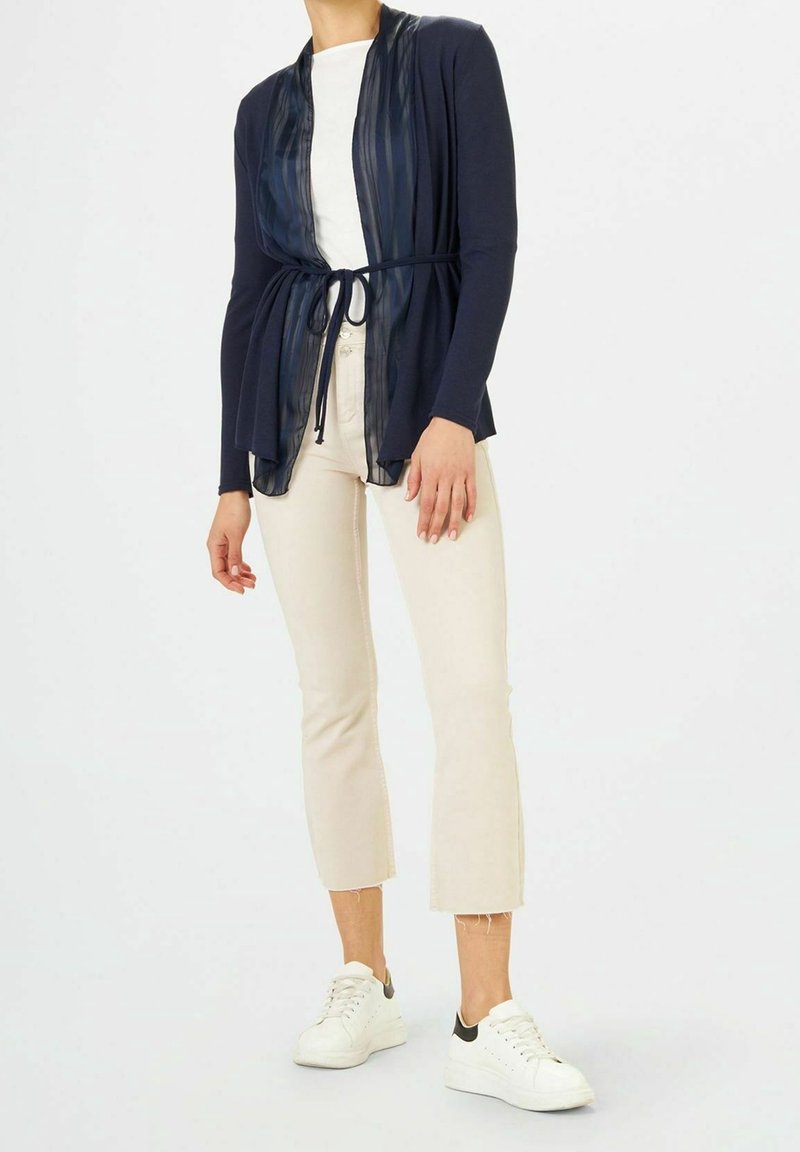 Femme portant un cardigan bleu marine ceinturé sur une chemise blanche, pantalon beige raccourci et baskets blanches sur fond uni.