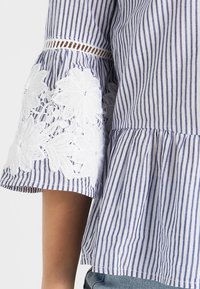 Blusa a righe blu e bianche con orlo peplum, caratterizzata da un dettaglio in pizzo ricamato bianco sulle maniche e tessuto strutturato.