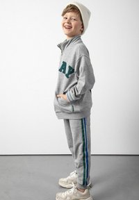 Szary, zapinany na zamek sweter z ciemnozielonym napisem, pasujące szare spodnie z niebieskimi paskami oraz biała czapka beanie, noszone z jasnymi trampkami.