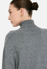 Grauer Rollkragensweater aus einem strukturierten Material mit subtilen Glitzerdetails. Verfügt über einen hohen Kragen und ein kleines Logo am hinteren Halsausschnitt.