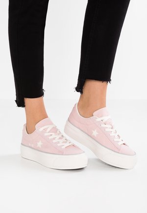 Osoba nosząca jasnoróżowe sneakersy na platformie z białymi sznurówkami i białym detalem w kształcie gwiazdy, w połączeniu z czarnymi spodniami z postrzępionym dołem.