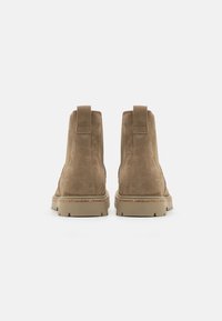 Botine din suede maro, cu un finisaj neted, vârfuri rotunjite, toc întărit, tălpi din cauciuc și mânere la spate pentru o îmbrăcare ușoară.
