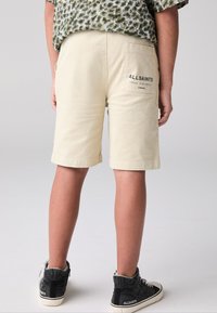 Beige bomullsshorts med uppvikta fållar, med en bakficka som har "ALLSAINTS"-logotypen. Bärs med svarta höga sneakers.