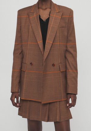 Osoba ubrana w brązową, kratkę marynarkę i pasującą plisowaną spódnicę na czarnej bluzce, stojąca na jednolitym szarym tle.