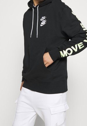 Mężczyzna ubrany w czarną bluzę z kapturem Nike z białymi sznureczkami i napisem „MOVE” na rękawie, połączoną z białymi spodniami typu cargo.
