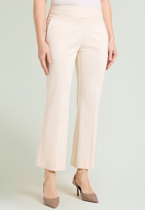 Frau trägt hoch taillierte, cremefarbene, gerade geschnittene Hosen, kombiniert mit taupefarbenen Slingback-Pumps mit spitzer Kappe, stehend vor einem schlichten Hintergrund.