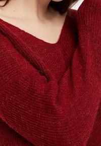 Czerwony sweter z dzianiny z głębokim dekoltem w kształcie litery V, z teksturowanej włóczki i widocznymi włóknami, podkreślający ciepło i miękkość.