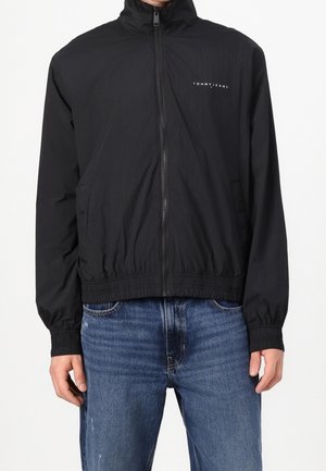 Veste légère noire zippée, avec poignets et ourlet élastiques, arborant un petit logo blanc "Tommy Jeans" sur la poitrine, portée avec un jean bleu.