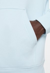 Felpa azzurra con tasca frontale. Tessuto morbido e polsini a coste. Una mano di carnagione scura poggiata nella tasca. Design semplice.