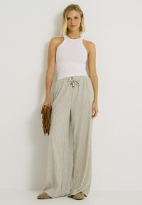 Mujer vestida con una blusa sin mangas blanca y pantalones de rayas beige de pierna ancha, sosteniendo un clutch marrón con flecos frente a un fondo blanco.