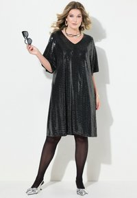 Schwarzes Pailletten-Kleid mit V-Ausschnitt, kurzen Ärmeln und lockerer Passform. Das Model trägt schwarze Strumpfhosen und Schuhe mit silbernen Akzenten und hält eine Sonnenbrille.