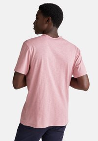 Joven con cabello corto y rizado, vistiendo una camiseta de mangas cortas de color rosa claro y pantalones oscuros, de espaldas contra un fondo blanco.