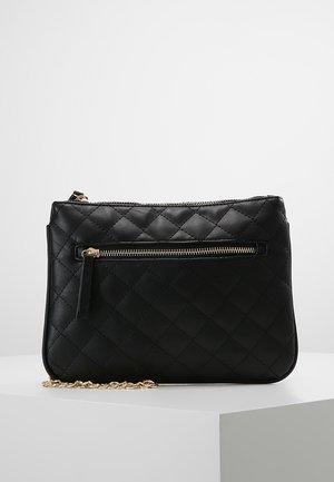 Czarna pikowana kopertówka ze skóry z złotą kieszenią na zamek z przodu i złotym łańcuchowym paskiem, spoczywająca na białym postumencie.