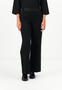 Schwarze weite Hosen aus strukturiertem Stoff. Mit einer hohen Taille und einem elastischen Bund mit Logo. Kombiniert mit glänzenden schwarzen Loafers.