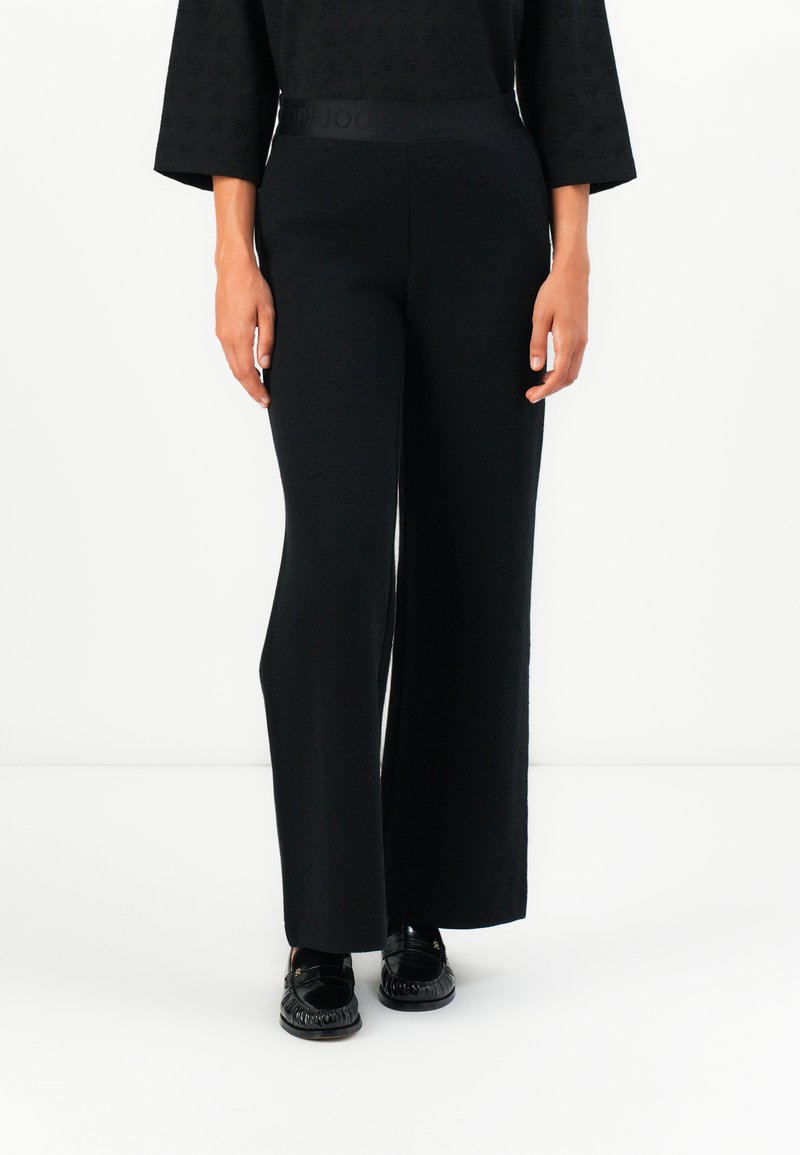 Schwarze weite Hosen aus strukturiertem Stoff. Mit einer hohen Taille und einem elastischen Bund mit Logo. Kombiniert mit glänzenden schwarzen Loafers.