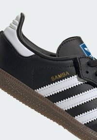 Čierne kožené tenisky s bielymi pruhmi a zlatým textom "SAMBA". Obsahujú gumenú podrážku s geometrickým vzorom a polstrovaným golierom.