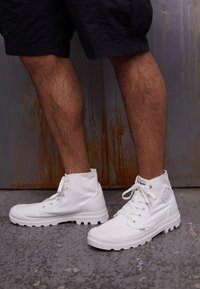 Sneakers bianchi a collo alto con una parte superiore in tessuto canvas testurizzato, punta rotonda e suola in gomma robusta. Presentano un'etichetta logo e lacci a contrasto.