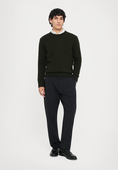 Pull en maille vert foncé avec des poignets côtelés, associé à un pantalon plissé noir et des chaussures habillées noires, porté par une personne se tenant contre un fond uni.