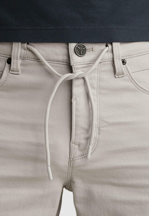 Pantalones beige claro con cordón ajustable atado en un lazo en la cintura sobre una camisa negra, destacando los detalles frontales de botones y bolsillo.