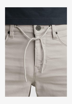 Pantalones beige claro con cordón ajustable atado en un lazo en la cintura sobre una camisa negra, destacando los detalles frontales de botones y bolsillo.