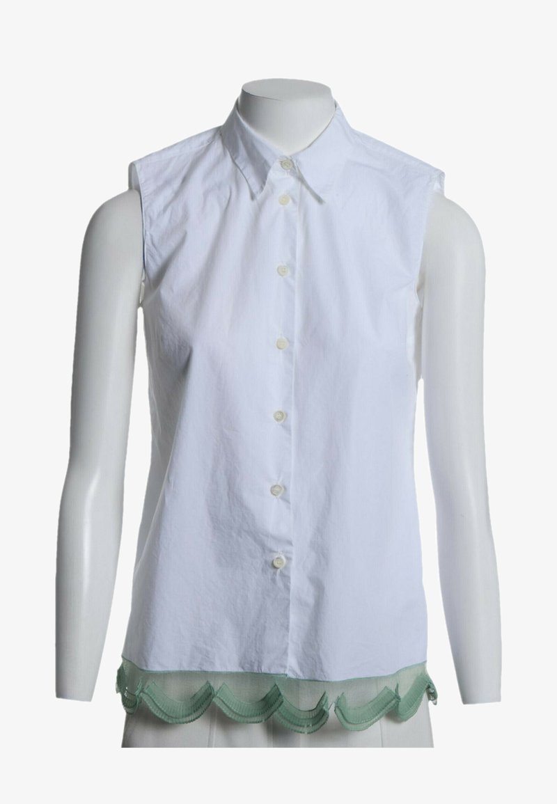 Chemise blanche sans manches à boutons avec col pointu et bordure verte festonnée au bas, présentée sur un torse de mannequin blanc.