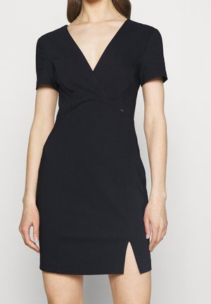 Vrouw die een korte zwarte jurk draagt met korte mouwen, V-hals, getailleerde taille en een voorste dijsplit aan één kant.