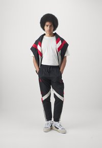 Černý sportovní teplákový oblek s červenými a bílými šikmými pruhy, který zahrnuje krátký rukáv na bundě a zúžené kalhoty, doplněný teniskami.