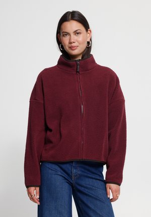 Femme portant une veste polaire bordeaux avec une fermeture éclair à l'avant et un jean bleu, debout devant un fond clair uni.
