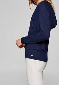 Dunkelblauer Pullover-Hoodie aus weichem Stoff, mit langen Ärmeln, Kordelzugkapuze und einem kleinen Logodetail am Saum, kombiniert mit weißen Hosen.