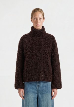 Femme portant une veste polaire texturée marron foncé avec un col montant et un jean bleu sur un fond clair uni.