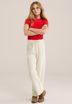 Noor tüdruk, kellel on pikad blondid juuksed ja prillid, seisab käed rüpes, kandes punast särki, triibulisi pükse ja pruune saapaid.