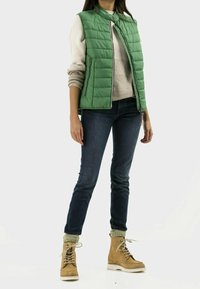 Gilet matelassé vert avec un col haut, porté sur un pull beige, associé à un jean foncé et des bottes à lacets beiges avec des chaussettes à accents verts.