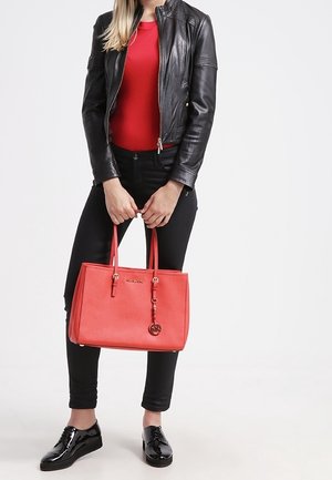 Sac à main en cuir rouge avec double anses, forme rectangulaire, surface texturée et accent de logo métallique. Porté par une personne en veste noire.