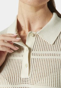 Pletené polo tričko v krémové barvě s texturovaným perforovaným designem; má límec a dva knoflíky. Ruka jemně spočívá na límci.