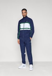 Chaqueta deportiva azul marino con rayas blancas y verdes, cierre frontal con cremallera, pantalones a juego, puños elásticos y detalles con el logo. Zapatillas deportivas blancas completan el conjunto.