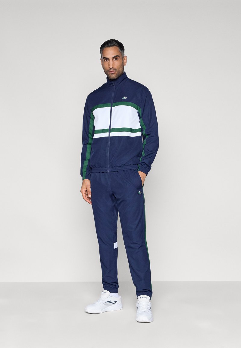 Chaqueta deportiva azul marino con rayas blancas y verdes, cierre frontal con cremallera, pantalones a juego, puños elásticos y detalles con el logo. Zapatillas deportivas blancas completan el conjunto.