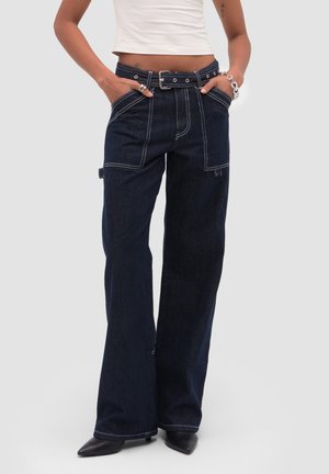 Osoba nosząca ciemne dżinsy o szerokich nogawkach z widocznym białym przeszyciem, szlufkami na pasek, przednimi i bocznymi kieszeniami, w połączeniu z czarnymi butami na spiczastym nosku.