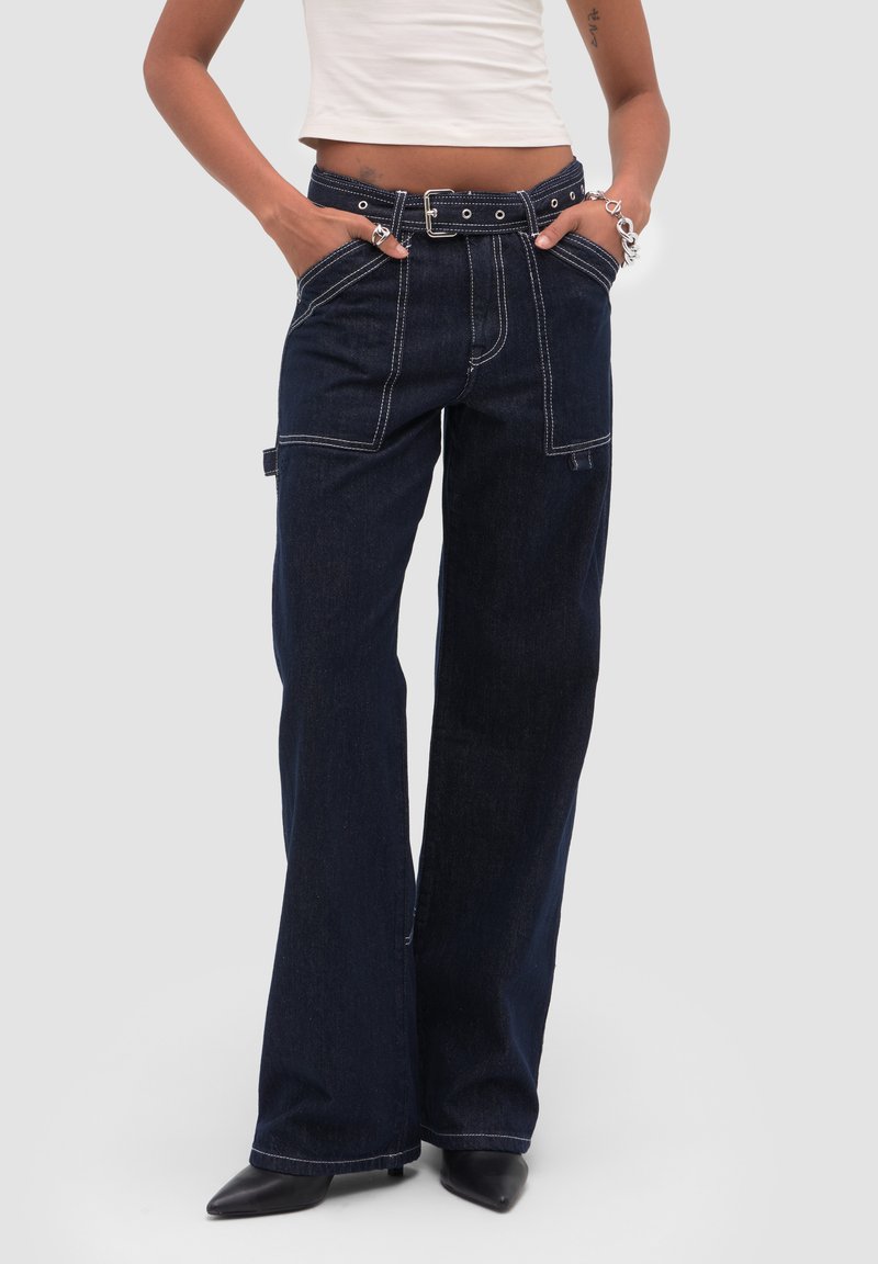 Personne portant un jean large foncé avec des coutures blanches visibles, des passants de ceinture, des poches avant et latérales, associé à des chaussures noires à bout pointu.