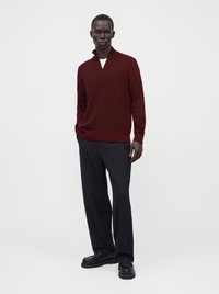Homme debout portant un pull bordeaux à fermeture éclair quart de zip, pantalon noir à jambes larges, chaussures noires, mains dans les poches sur un fond blanc uni.